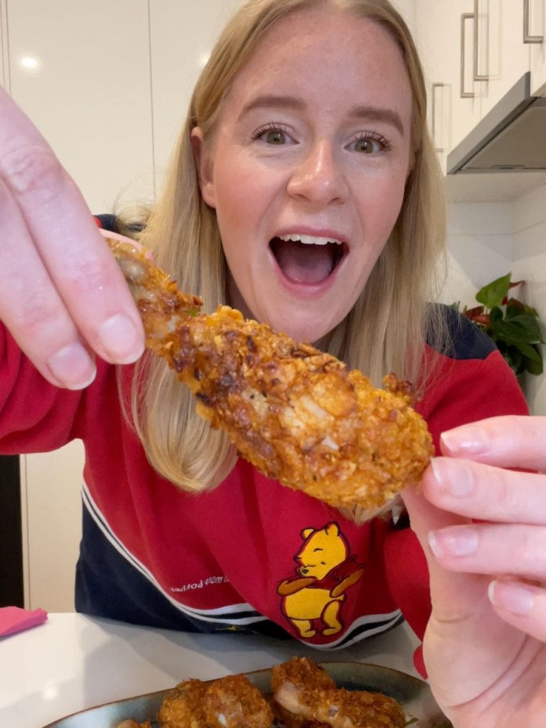 Blonde woman smiling while holding up a crispy chicken drumstick, golden and perfectly breaded