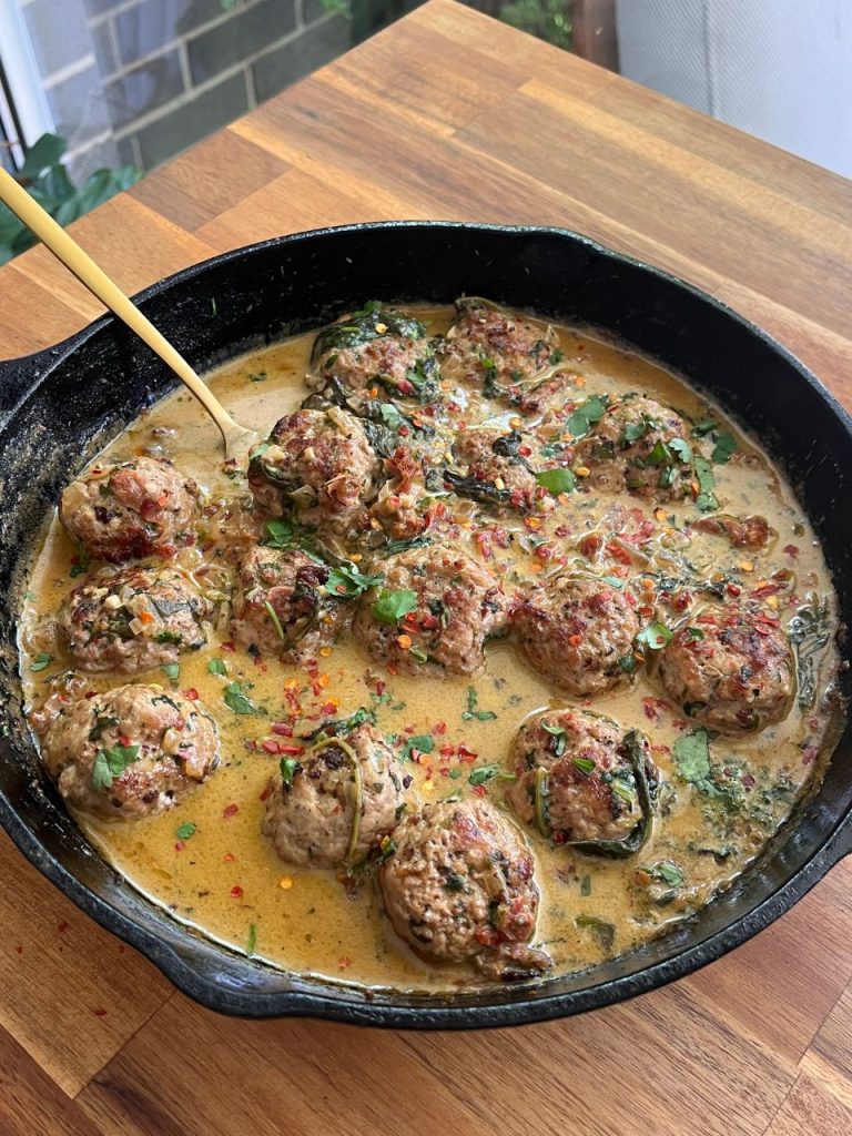 Golden pork meatballs coated in creamy sauce in a black skillet, with a brass spoon resting inside