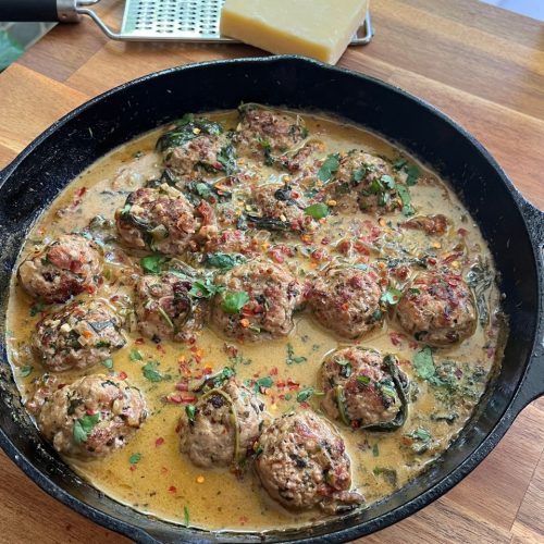 Golden pork meatballs coated in creamy sauce in a black skillet, with a brass spoon resting inside and parmesan cheese and a grater blurred in the background