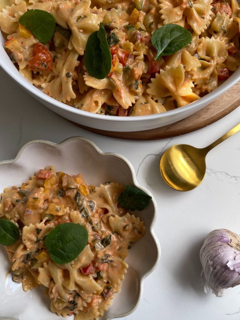 Creamy loaded vegetable pasta served in a large white serving bowl, with a smaller scalloped-edge bowl beside it and a brass spoon resting nearby.
