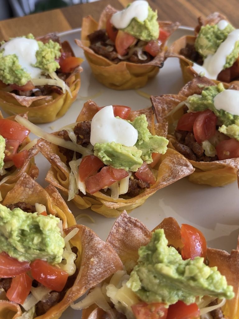 Finished taco cups topped with diced cherry tomatoes, smashed avocado, and a dollop of sour cream