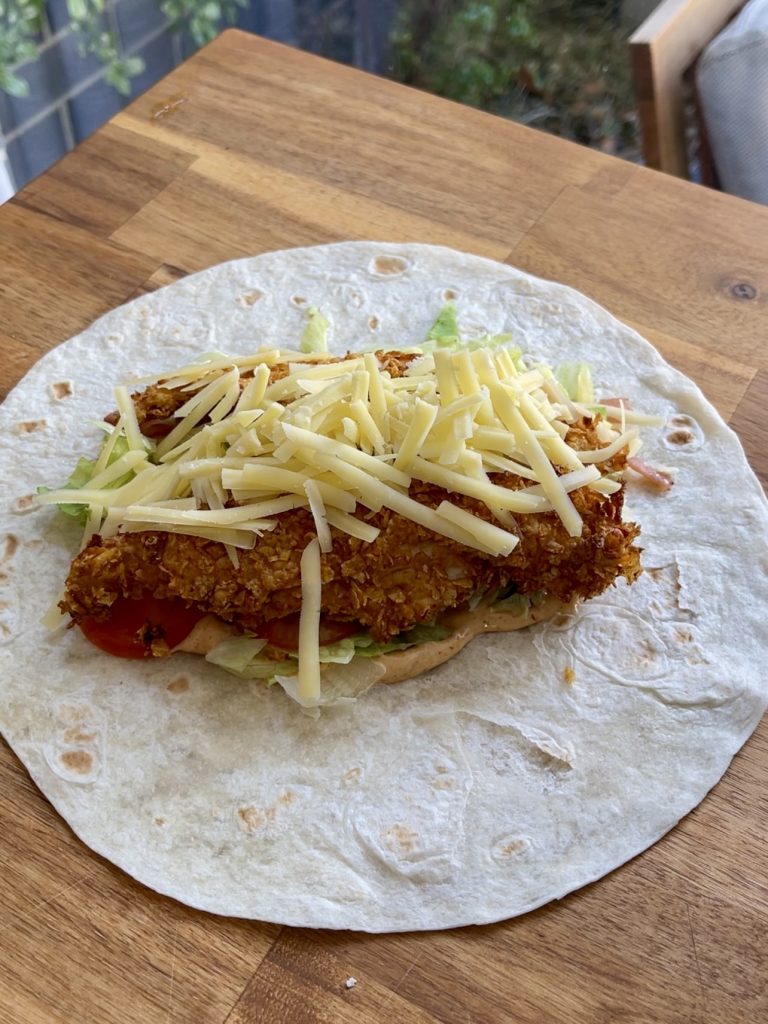 Tortilla laid flat with chipotle mayo spread in the centre, layered with lettuce, tomato, bacon, cheese, and crispy chicken strips