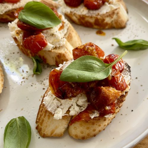 Roast tomato and whipped feta toast served on a ceramic plate, topped with basil leaves and cracked pepper